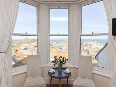 A bay window with two upholstered chairs and a round table with a vase of roses and cups overlooking rooftops and sea at Whistler in St Ives