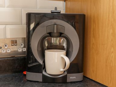 A coffee machine with a white mug on a kitchen countertop next to a wooden cabinet and tiled wall at Whistler in St Ives