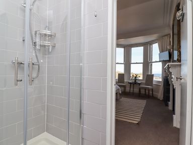 A bathroom with a glass shower enclosure and a bedroom with a striped rug two chairs and a table near bay windows at Whistler in St Ives
