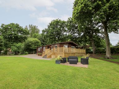 A cabin with a wooden deck and outdoor seating area on a grassy lawn surrounded by trees at The Log Cabin in Newnham Bridge near Tenbury Wells