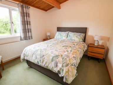 A bedroom with a double bed with leaf patterned bedding two wooden bedside tables with lamps and a window with checkered curtains at The Log Cabin in Newnham Bridge near Tenbury Wells