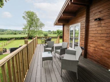 A wooden deck with outdoor chairs and tables overlooking a grassy area and trees at The Log Cabin in Newnham Bridge near Tenbury Wells