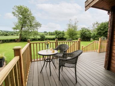 An outdoor wooden deck with two wicker chairs and a table with flowers overlooking a grassy yard at The Log Cabin in Newnham Bridge near Tenbury Wells