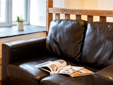 A black leather sofa with magazines and reading glasses on the seat near a window with a small potted plant at Stable Cottage in Port Isaac