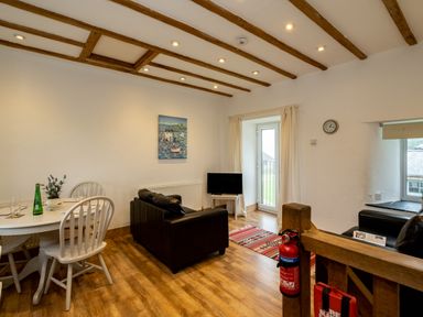 A living room with a black sofa a small television a round white dining table with chairs and wooden ceiling beams at Stable Cottage in Port Isaac