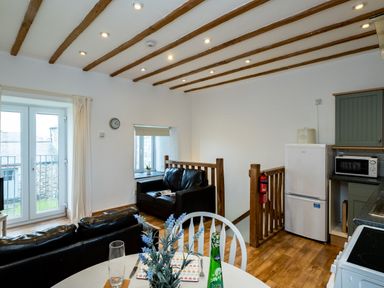 A kitchen and living area with black leather sofas a round dining table with chairs wooden ceiling beams and a small staircase at Stable Cottage in Port Isaac