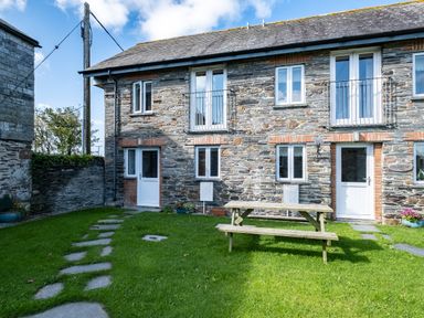 A stone house with multiple white doors and windows a wooden picnic table and green lawn in front at Stable Cottage in Port Isaac