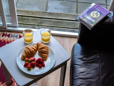 A small table with two croissants strawberries raspberries and two glasses of orange juice next to a leather chair with books on the armrest at Atlantic Lodge in Port Isaac