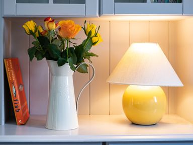 A white pitcher with yellow and orange roses next to a yellow lamp and an orange book on a white shelf at Atlantic Lodge in Port Isaac