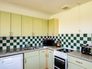 A kitchen with green cabinets green and white tiled backsplash a dishwasher a sink a microwave a kettle an oven and a toaster at Atlantic Lodge in Port Isaac