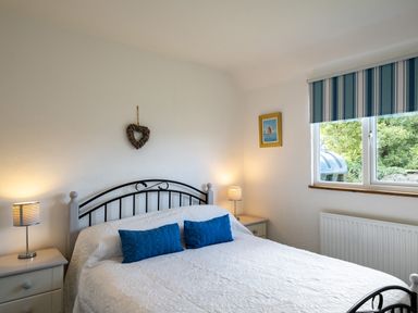 A bedroom with a double bed two blue pillows two bedside tables with lamps a window with striped blinds and a heart decoration on the wall at Atlantic Lodge in Port Isaac
