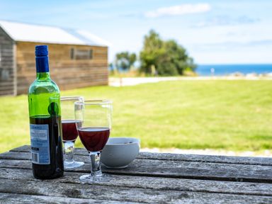 A wooden picnic table with a bottle of red wine, two glasses with red wine, and a bowl outdoors with grass and a wooden shed in the background at Atlantic Lodge in Port Isaac