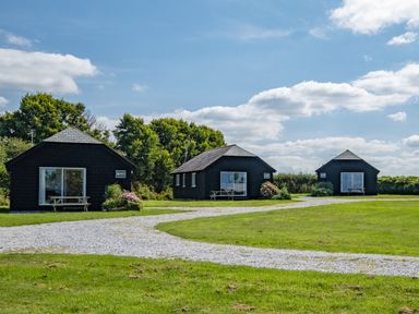 Three black wooden cabins with picnic tables in front on a grassy area with a gravel path and trees in the background at Atlantic Lodge in Port Isaac