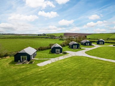Five small houses with patios on a grassy field with a gravel path in a rural landscape at Atlantic Lodge in Port Isaac