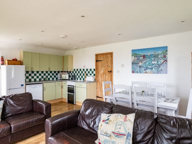 An open living space with a kitchen area including green cabinets and checkerboard backsplash a dining table with white chairs and dark brown leather sofas in the foreground at Gull Lodge in Port Isaac