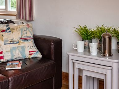 A leather couch with a nautical themed pillow and glasses on a book next to nested white tables with mugs a metal coffee pot and plants at Gull Lodge in Port Isaac