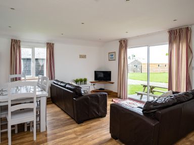 A living room with two brown leather sofas a white dining table with chairs a small TV on a corner shelf and sliding glass doors overlooking a grassy yard at Gull Lodge in Port Isaac