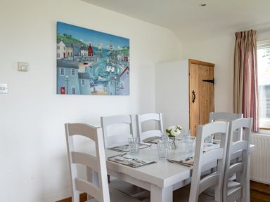A dining area with a white table and chairs set with placemats and glasses and a painting on the wall at Gull Lodge in Port Isaac
