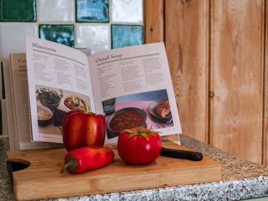 A cutting board with a red bell pepper a tomato and a red chili pepper in a kitchen with a recipe book open at Minestrone and Oxtail Soup recipes