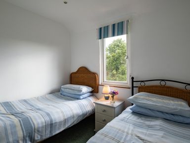 A bedroom with two single beds a window with striped blind and a bedside table with a lamp and flowers at Gull Lodge in Port Isaac