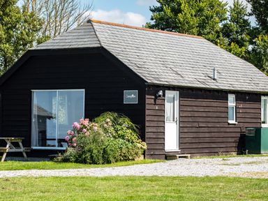 A black wooden cottage with a gray tiled roof flowers in front and a picnic bench on the side at Campion Lodge in Port Isaac