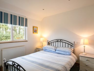 A bedroom with a double bed with striped bedding two bedside tables with lamps a window with striped blinds and a framed picture on the wall at Campion Lodge in Port Isaac