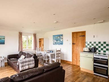 A living and dining area with two brown leather sofas a white dining table with chairs a kitchen corner with appliances and wooden doors at Campion Lodge in Port Isaac