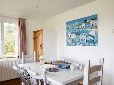A dining room with a white table and chairs puzzle pieces and two mugs on table and a window with curtains at Campion Lodge Port Isaac