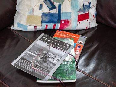 A pair of reading glasses on top of Port Isaac village trail and Newquay Padstow maps on a leather couch with a cushion