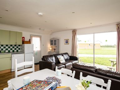 A living room with brown leather sofas a white dining table with chairs kitchen cabinets and a sliding glass door showing an outdoor area at Campion Lodge in Port Isaac