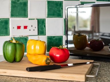 A kitchen cutting board with a knife and different colored peppers in front of a tiled wall and microwave at Campion Lodge in Port Isaac