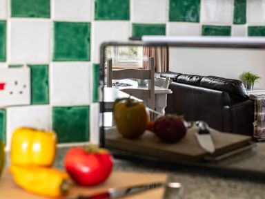 A kitchen counter with peppers and a cutting board in front of green and white tiles reflecting a dining area with chairs and a living room with a sofa and plant at Campion Lodge in Port Isaac