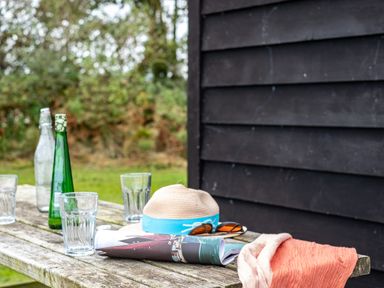 An outdoor wooden table with drinking glasses bottles a hat sunglasses a magazine and a cloth near a black wall at Campion Lodge in Port Isaac
