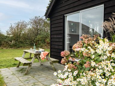 A wooden picnic table with glasses, bottle, and a hat on a patio outside a house with dark siding and a sliding glass door at Campion Lodge in Port Isaac
