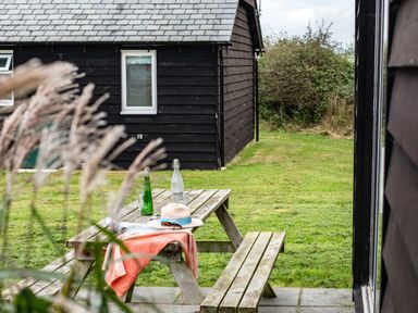 A wooden picnic table with bottles glasses a hat and a cloth in a grassy yard near black wooden buildings at Campion Lodge in Port Isaac