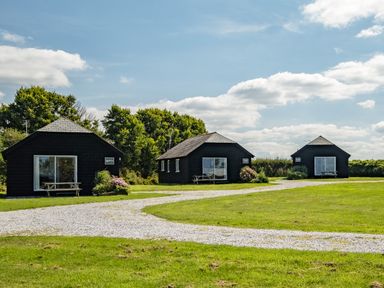 Three black wooden cabins with picnic tables on a grassy lawn and a gravel path under a blue sky with clouds