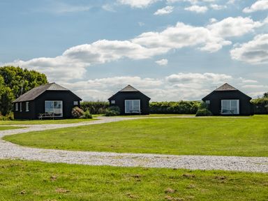 Three black small cottages on a grassy field with a gravel path under a cloudy sky at Campion Lodge in Port Isaac