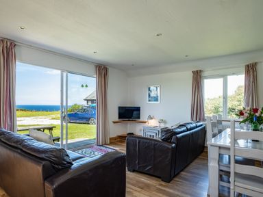 A living room with two dark sofas a corner TV table a white dining table with chairs and a large window with a sea view at Harbour Lodge in Port Isaac