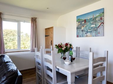 A dining room with a white dining table and chairs a vase with flowers two glasses of wine a bottle and a bowl on the table a window with curtains and a painting on the wall at Harbour Lodge in Port Isaac