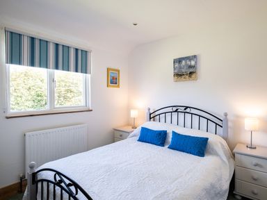 A bedroom with a double bed with white bedding and two blue pillows two bedside tables with lamps and a window with striped blinds at Harbour Lodge in Port Isaac