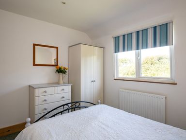 A bedroom with a white bedspread a white dresser with yellow flowers a white wardrobe and a window with striped blinds at Harbour Lodge in Port Isaac