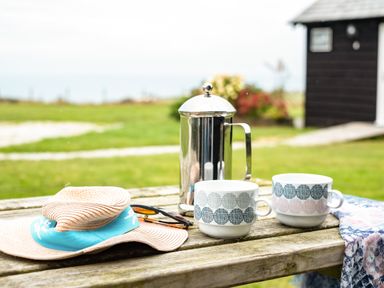 An outdoor wooden table with a straw hat, sunglasses, a metal coffee press, and two patterned mugs