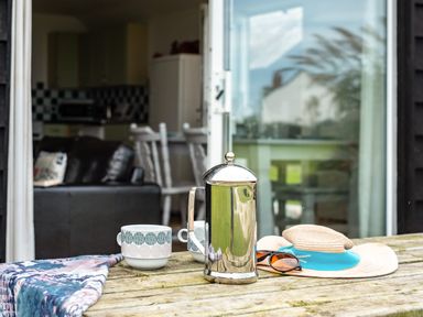 A wooden table with a metal coffee press two mugs sunglasses and a sun hat outside a room with open doors at Puffin Lodge in Port Isaac