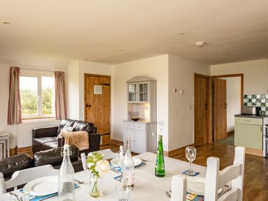A living room and dining area with a white table set with plates and bottles a gray cabinet two wooden doors and a kitchen area visible at Puffin Lodge in Port Isaac