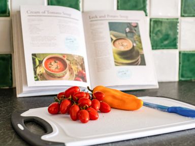 A kitchen counter with a cutting board holding cherry tomatoes an orange pepper and a blue knife with a cookbook in the background