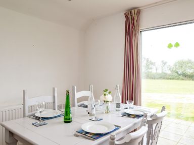 A dining table set with plates glasses bottles and flowers near a large window with striped curtains at Puffin Lodge in Port Isaac