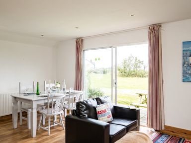 A dining area with a white table set with plates and glasses next to a black sofa and sliding glass doors showing a grassy yard at Puffin Lodge in Port Isaac
