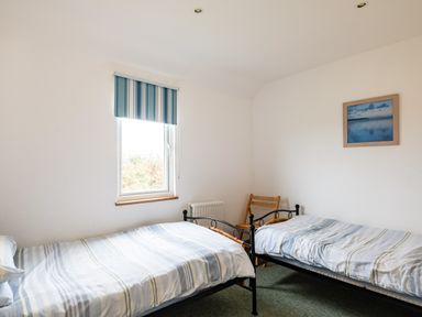 A bedroom with two single beds, a wooden chair, a window with striped blinds, and a framed picture on the wall at Dolphin Lodge in Port Isaac