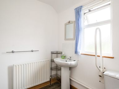 A bathroom with a sink, mirror, radiator, metal shelf, window with blue curtain, and a toilet with a support rail at Dolphin Lodge in Port Isaac