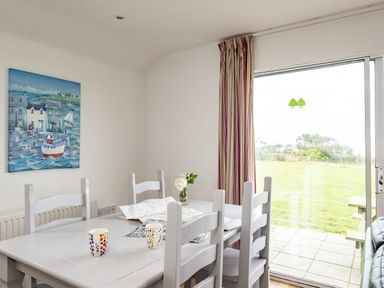 A dining area with a white table and chairs with a map and mugs on the table a glass door leading to a garden with a picnic table and a painting of a seaside scene on the wall at Dolphin Lodge in Port Isaac